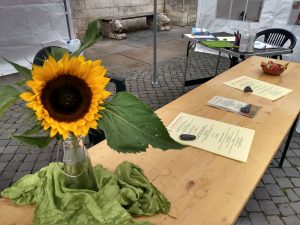 Auf einem Bierzelt-Tisch liegen Flyer und Handzettel zu Schreibwerkstätten. Vorn auf dem Tisch steht eine Sonnenblume in einer Vase, die auf einem grünen Tuch steht. Der Tisch befindet sich unter einem Pavillon.